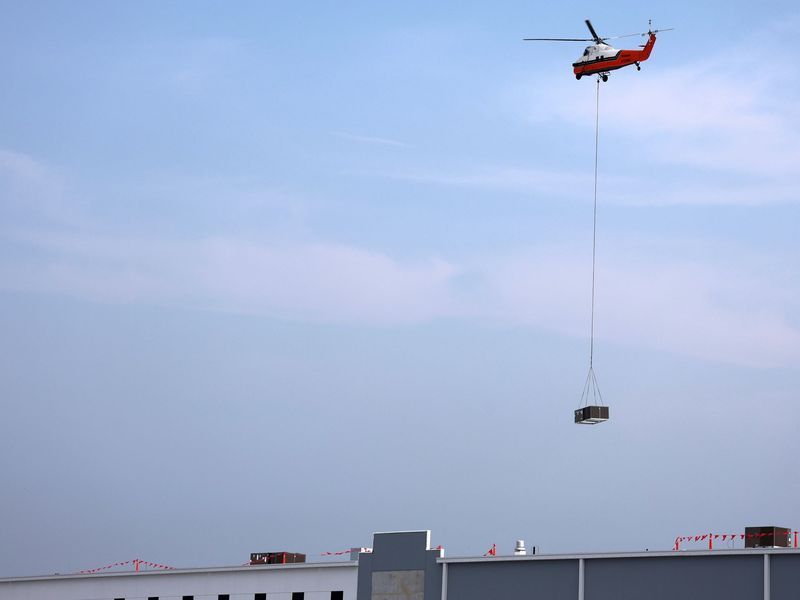 A red and white helicopter hovers in a clear sky, using a long cable to lower a container onto a building rooftop.