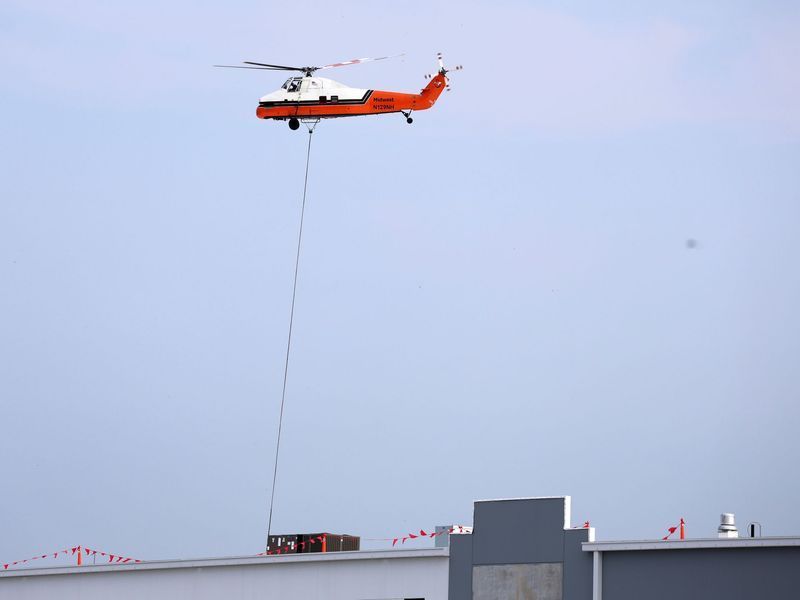 An orange and white helicopter flying over a building while carrying a load on a long cable.