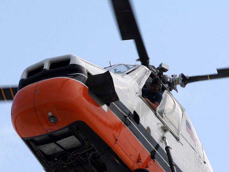 A white and orange helicopter in flight with a crew member visible in the cockpit against a clear sky.