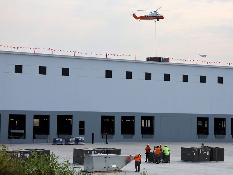 A helicopter lowers an HVAC unit onto a large industrial building roof while workers wait on the concrete ground below.