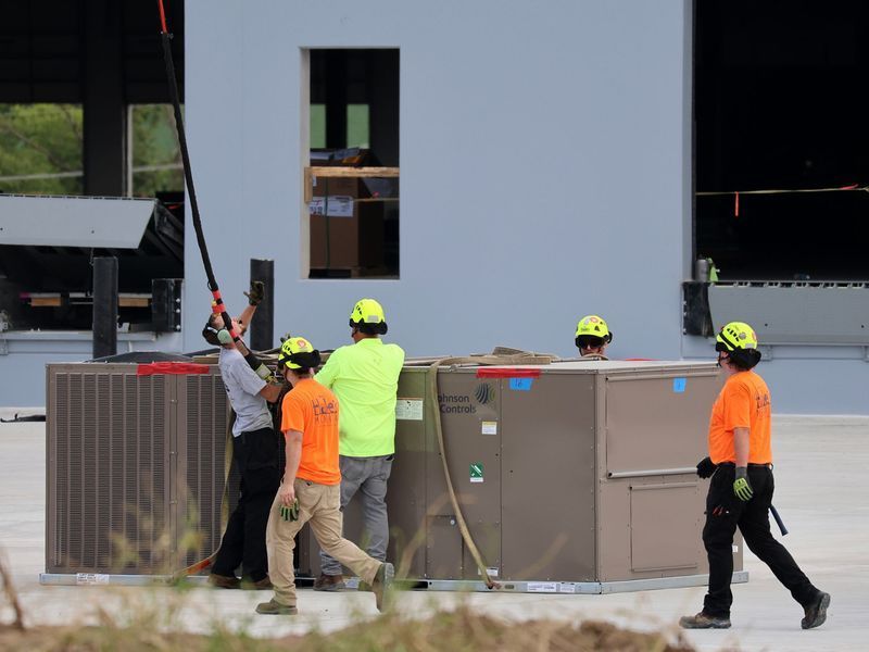 Construction workers in hard hats and safety gear move a large industrial HVAC unit on a building rooftop.