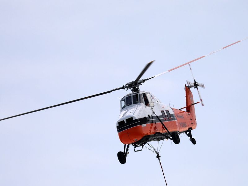 An orange and white helicopter flying against a pale sky, with a long cable hanging from its underside.