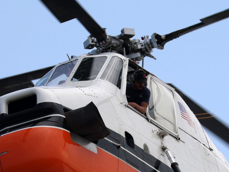 Close-up of a white and orange helicopter pilot leaning out of the cockpit against a clear blue sky.
