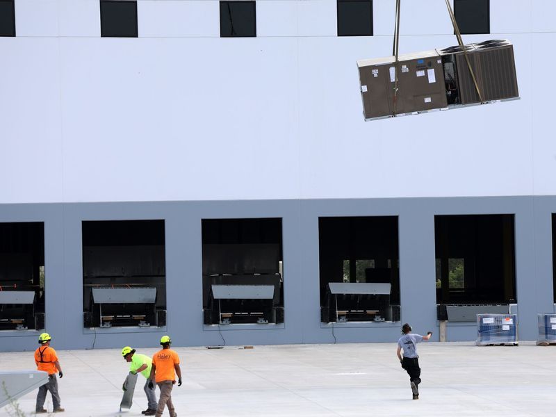 Construction workers watch as a crane lifts a large industrial HVAC unit toward the roof of a gray warehouse building.