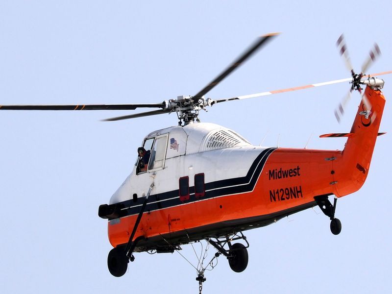 A white and orange Midwest helicopter in flight against a clear blue sky, carrying a load from a cable underneath.