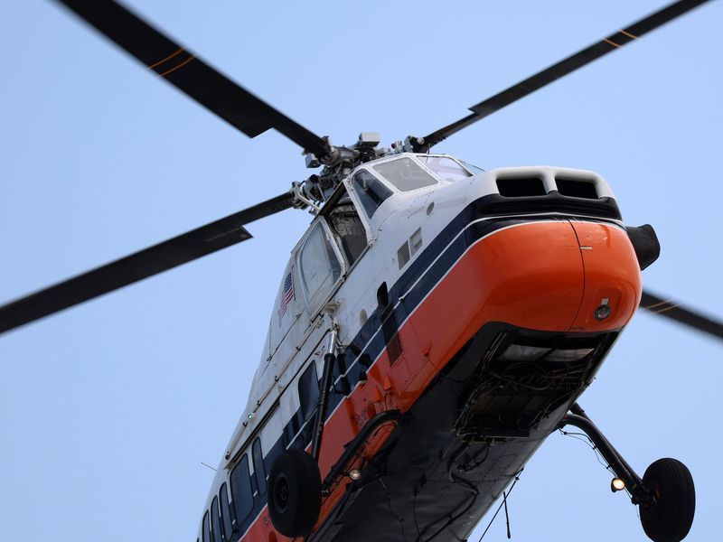 A white and orange helicopter in flight against a clear blue sky, viewed from a low angle.