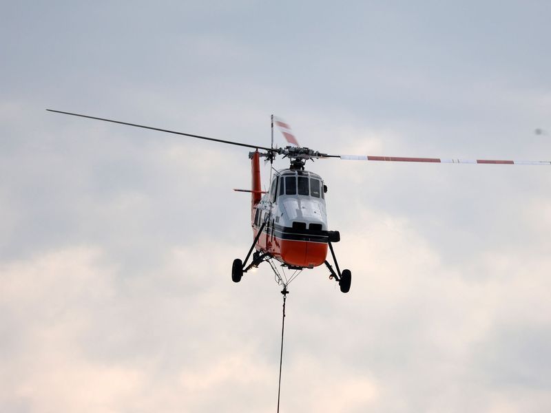 An orange and white helicopter in flight against a cloudy sky with a cable hanging from its underside.
