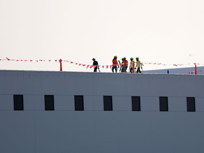 Several construction workers in high-visibility gear walk along the edge of a flat-roofed building marked by orange flags.