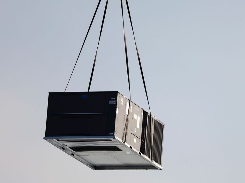 A large, dark HVAC rooftop unit is suspended in the air by straps, being lifted by a crane against a clear sky.