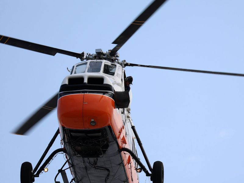 A white and orange rescue helicopter flying against a clear blue sky, viewed from a low angle.
