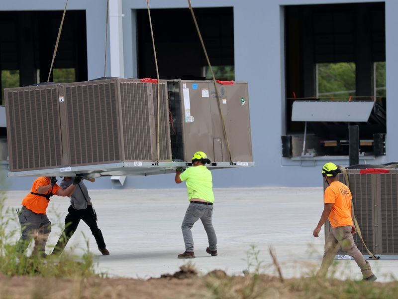 Four workers in high-visibility gear guide a large HVAC unit suspended by cables outside a commercial building.