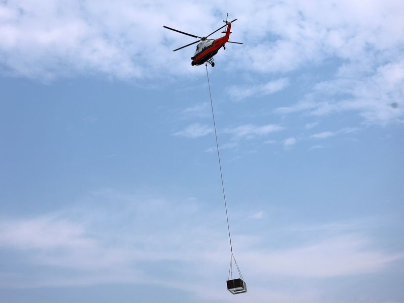 A red and white helicopter flies in a blue sky, carrying a black container suspended by a long cable.