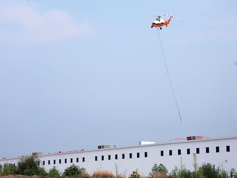 A white and orange helicopter hovers over a long, white building, carrying a long cable hanging from its underside.