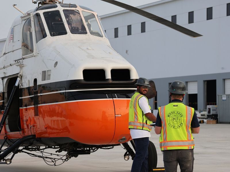 Two workers in high-visibility vests and hard hats stand next to an orange and white helicopter on a tarmac.