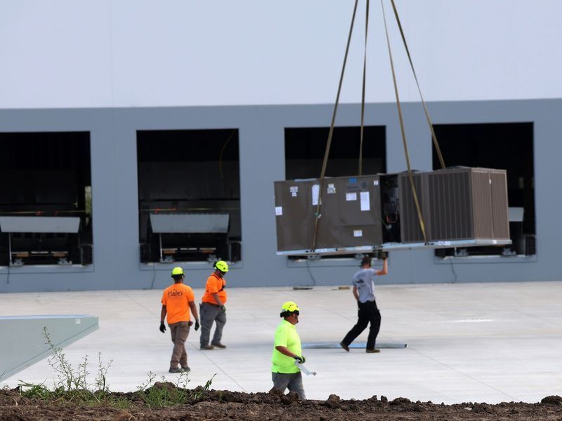 Four workers wearing high-visibility shirts oversee the crane installation of an HVAC unit on a building rooftop.