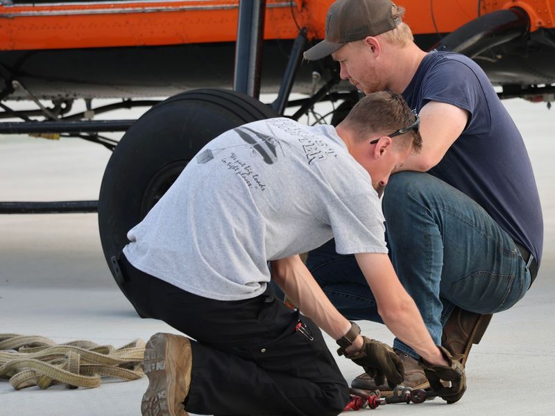 Two people in work clothes kneeling to repair the landing gear of an orange aircraft on a light-colored surface.