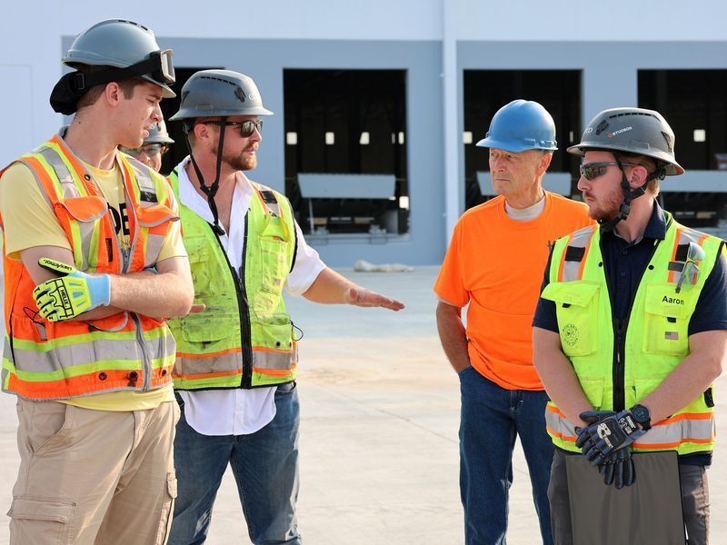 Four people in hard hats and safety vests stand outdoors, engaged in a discussion on a construction site.