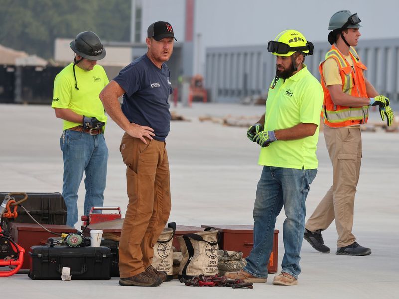Four construction workers in safety gear and hard hats stand on a flat, open job site with equipment cases on the ground.