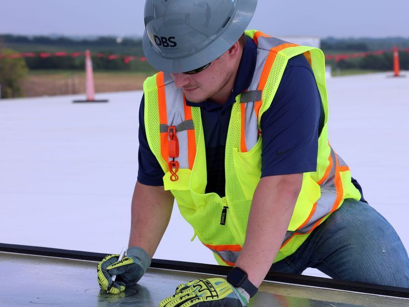 A construction worker in a high-visibility vest and hard hat works on a large, flat, white roof surface.