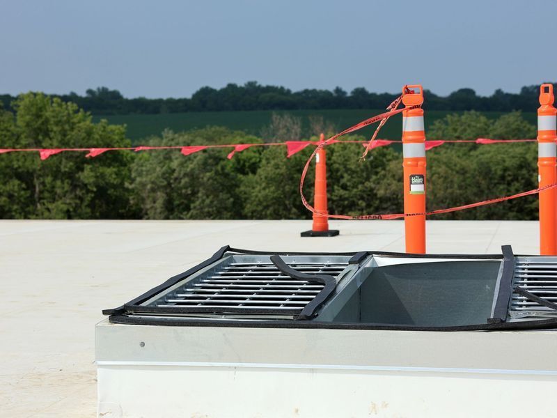 A roof access hatch with a safety grate, surrounded by orange traffic cones and caution tape against a rural background.