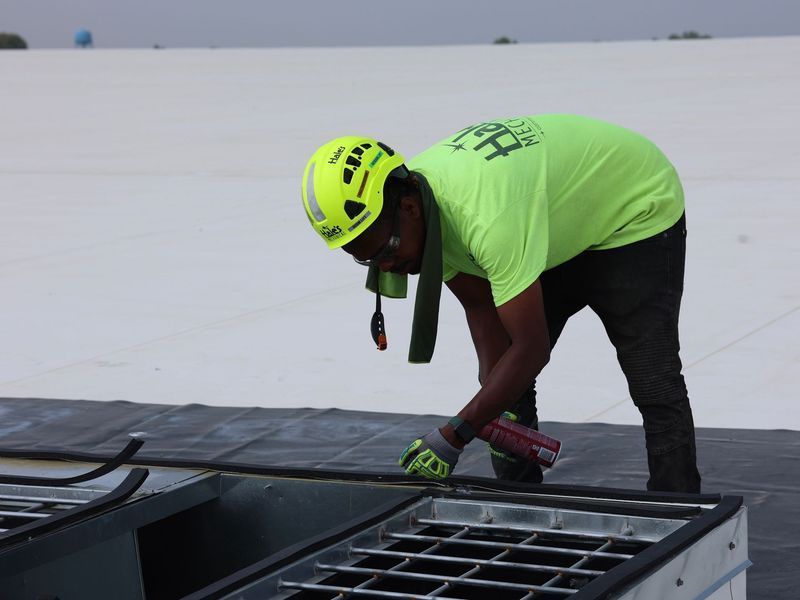 A worker in a neon yellow hard hat and shirt seals a seam around a rooftop vent on a white surface.