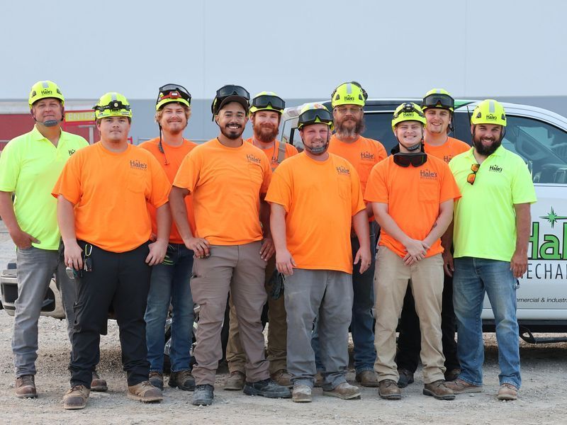A group of 11 workers in safety gear and uniform shirts posing in front of a company truck at an outdoor job site.
