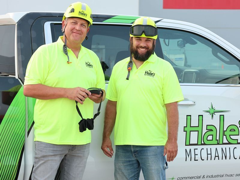 Two workers in lime-green uniform shirts and hard hats stand smiling in front of a branded Hale's Mechanical service truck.