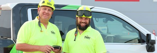 Two smiling individuals in neon-yellow work shirts and hard hats stand in front of a white service vehicle.