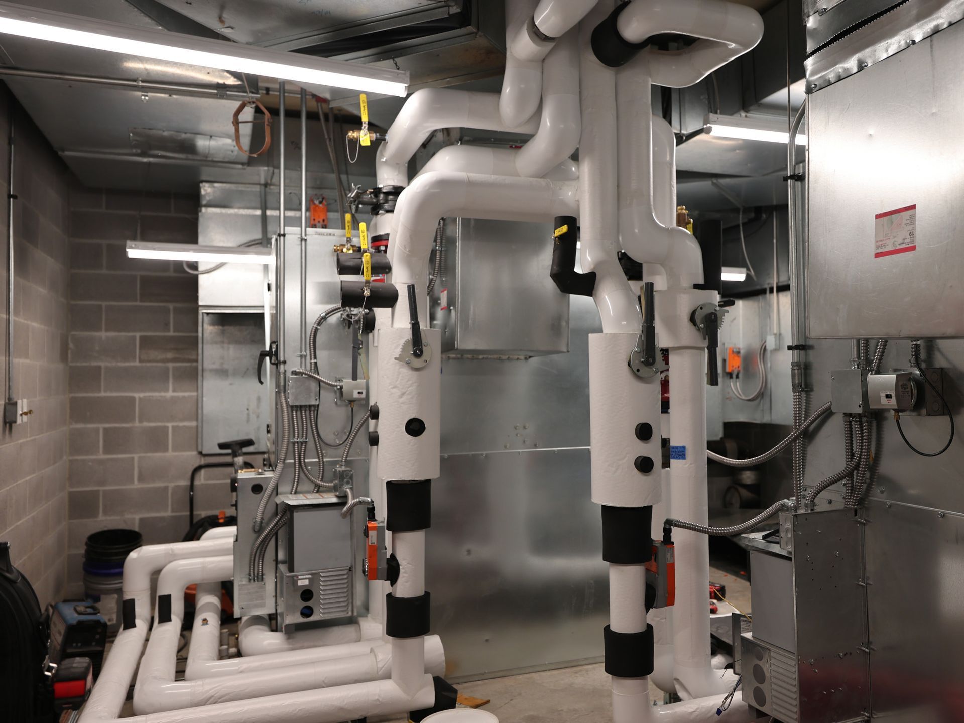 Industrial HVAC utility room featuring insulated white piping connected to metal equipment against a cinder block wall.