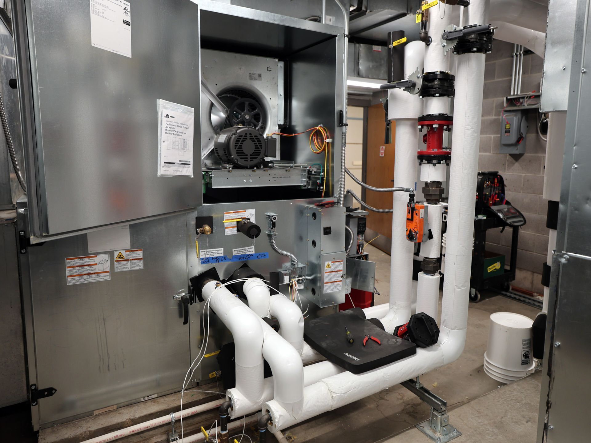 Industrial HVAC unit with insulated piping, valves, and a motor housed in a metal cabinet in a utility room.