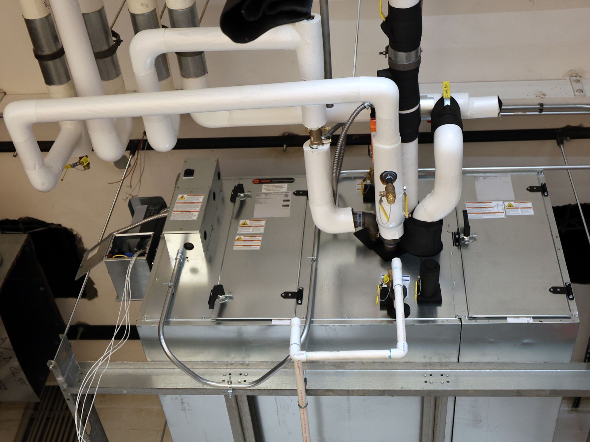 A view of a commercial HVAC air handling unit with white insulated pipes hanging from the ceiling in a mechanical room.