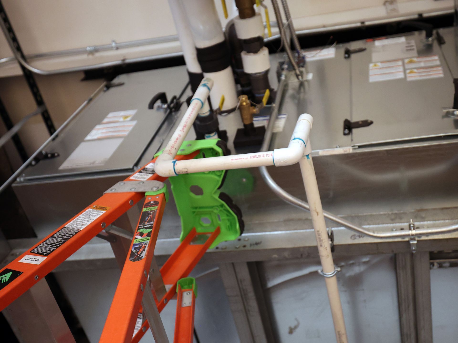 An orange stepladder placed next to white PVC pipes and industrial HVAC equipment inside a utility room.