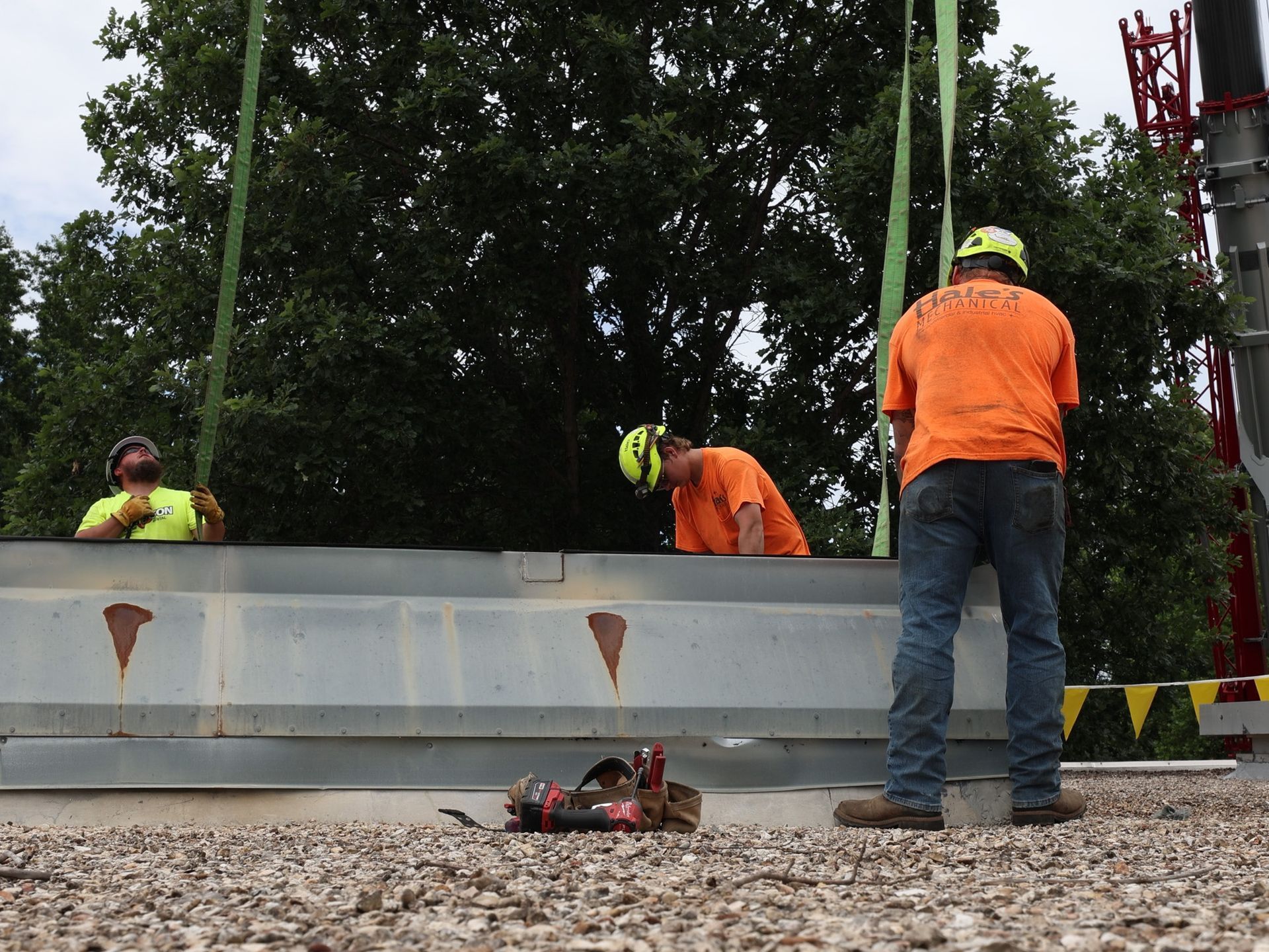 Three construction workers in hard hats and safety gear position a large metal beam suspended by a crane on a gravel roof.