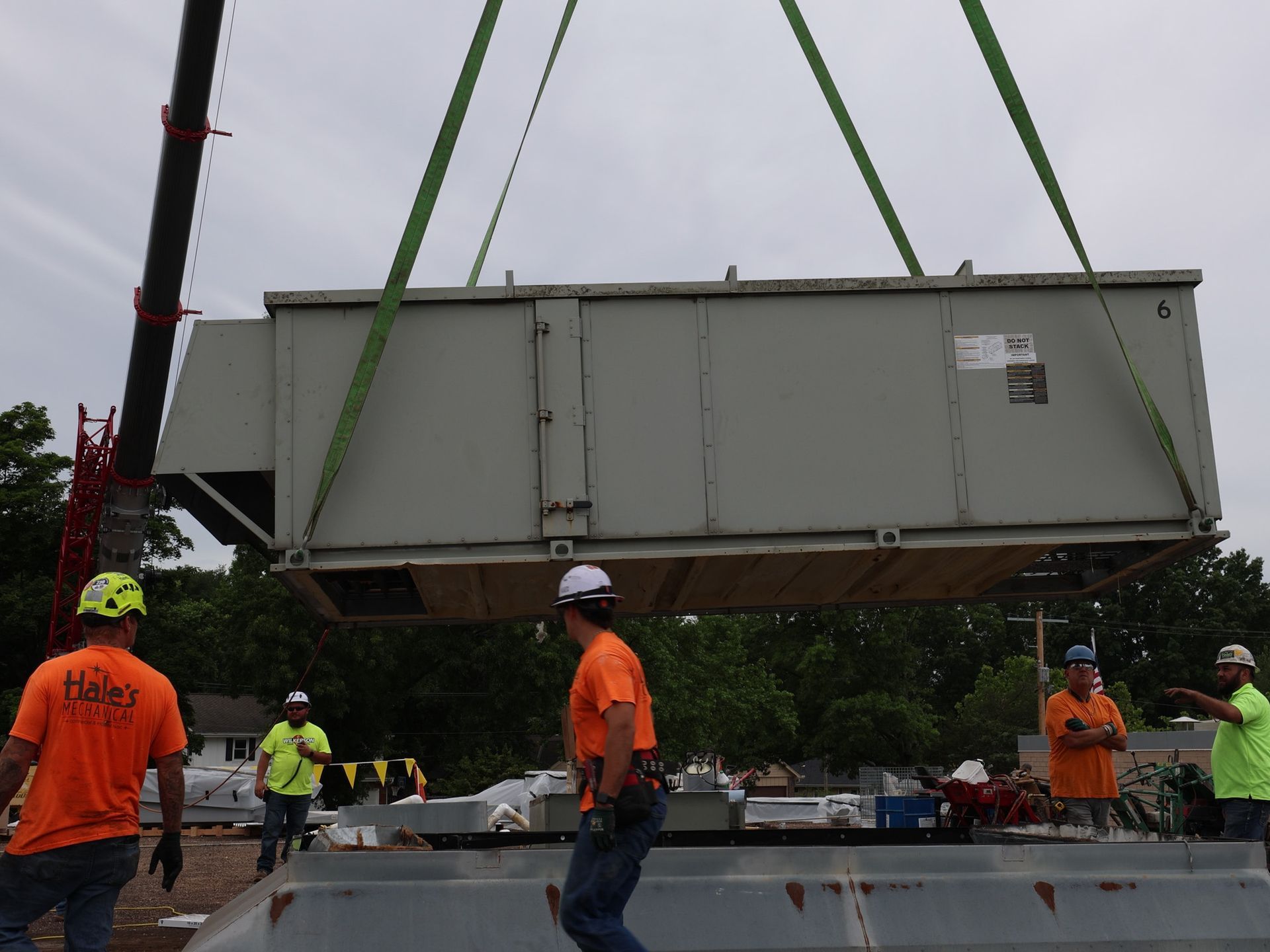 A crane lowers a large gray HVAC unit toward workers on a construction site.