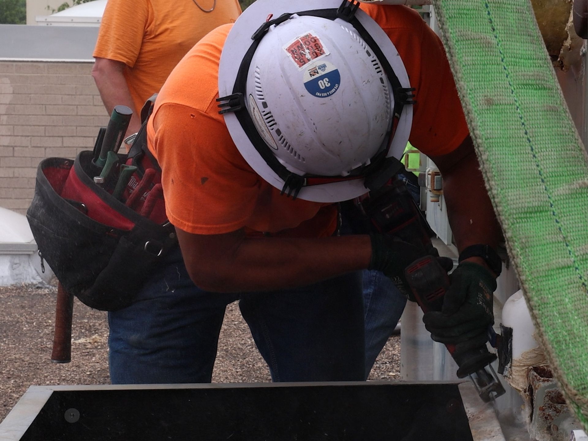 A construction worker in an orange shirt and white hard hat uses a reciprocating saw on a metal frame.