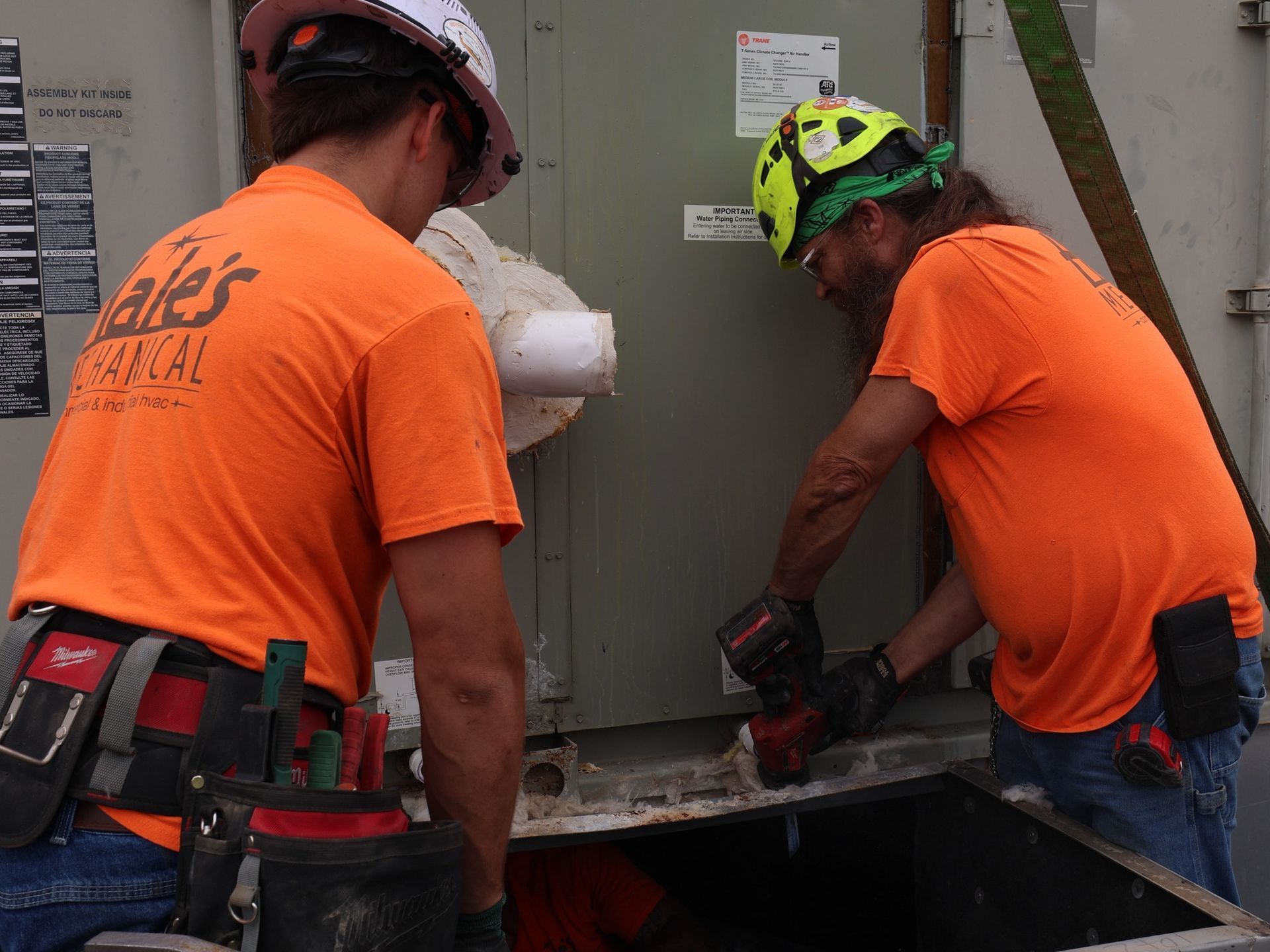 Two workers in orange shirts and hard hats use tools to install equipment near a large, grey industrial cabinet.