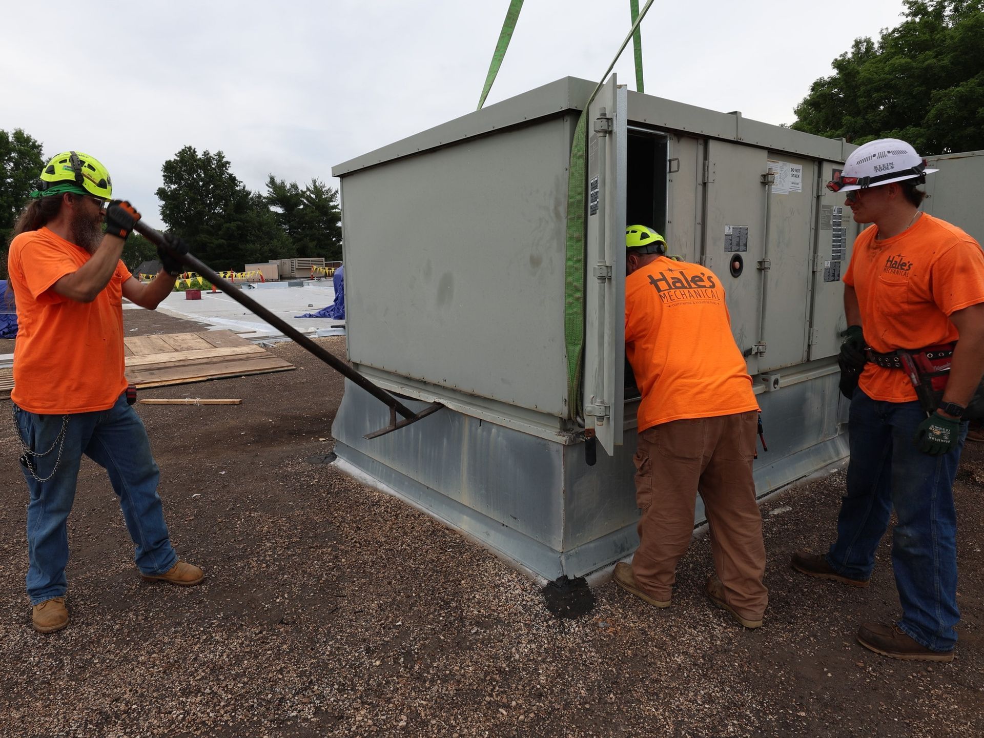 Three workers in orange shirts and safety gear use a pry bar to adjust a large, metal electrical transformer box outdoors.