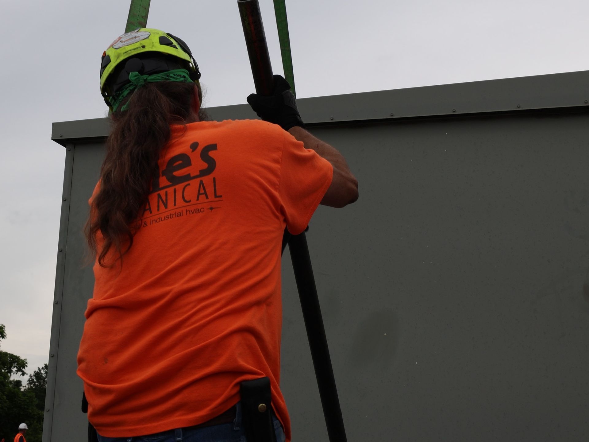 A worker in an orange t-shirt and safety helmet stabilizes a suspended load attached to a crane at a construction site.