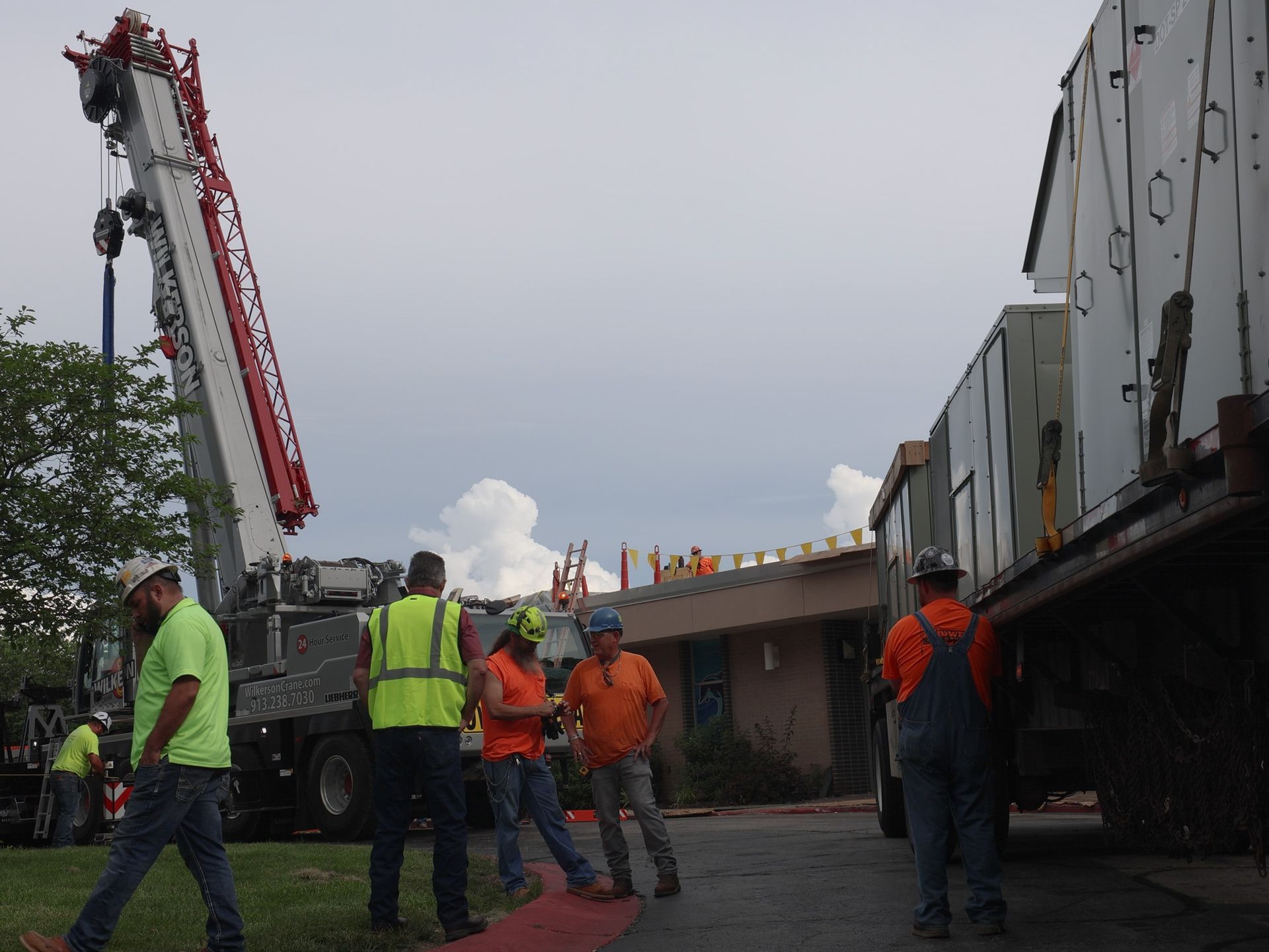 A group of workers in high-visibility gear stand outside a building next to a large mobile crane and a flatbed truck.