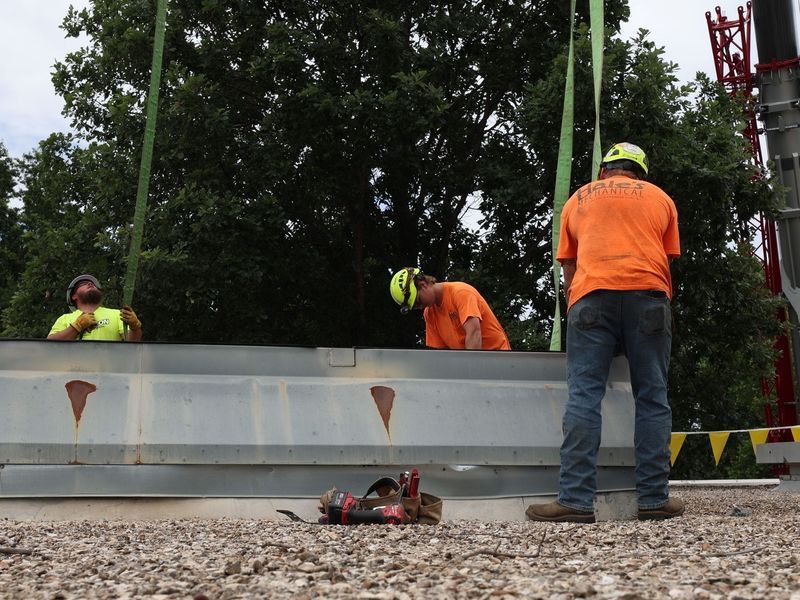 Three construction workers in hard hats and safety gear stand outdoors, guiding a large metal beam suspended by a crane.