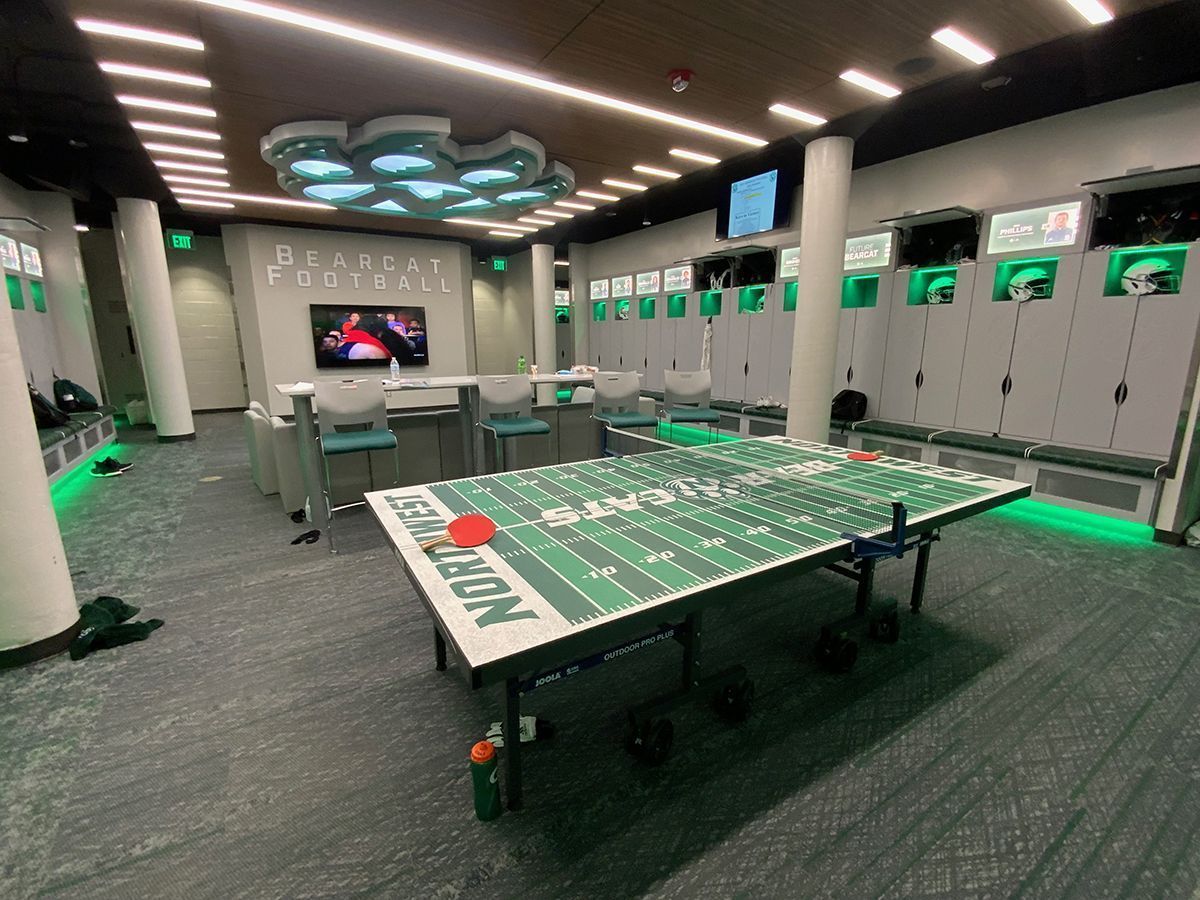 Locker room with a central ping-pong table featuring a green football field design, sleek lockers, and neon lighting.