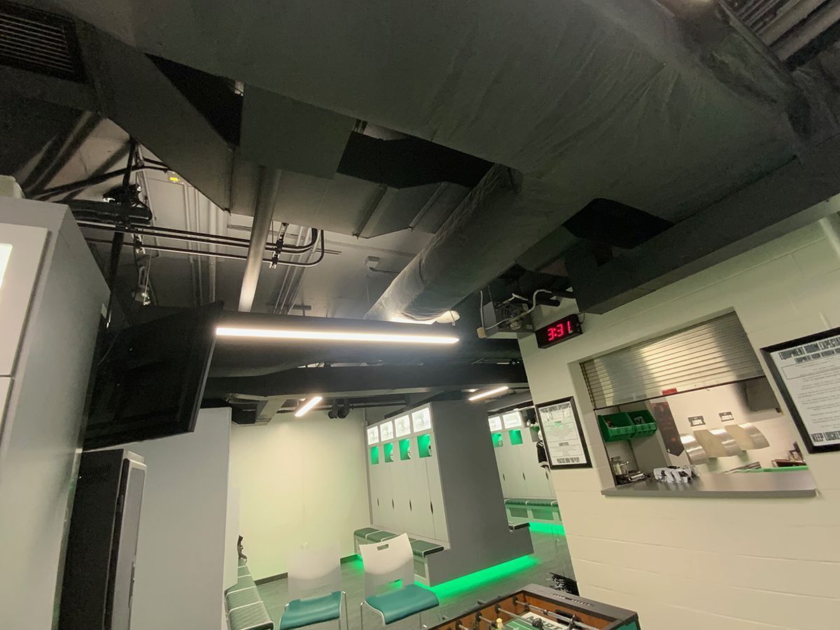 A modern office break room with exposed ductwork, fluorescent lighting, lockers, and a view into a kitchen area.