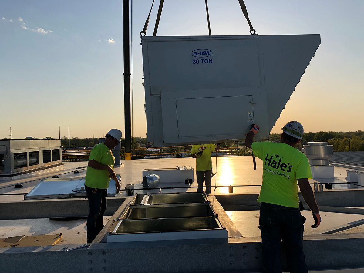 Workers in neon shirts guide a large white industrial HVAC unit being lowered by a crane onto a building rooftop at sunset.
