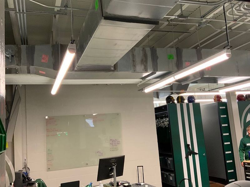 An office space with a whiteboard, a desk with a monitor, hanging lights, exposed ductwork, and green locker-style storage.