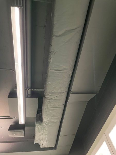 A view looking up at a ceiling with a long rectangular HVAC duct covered in silver insulation next to a linear light.