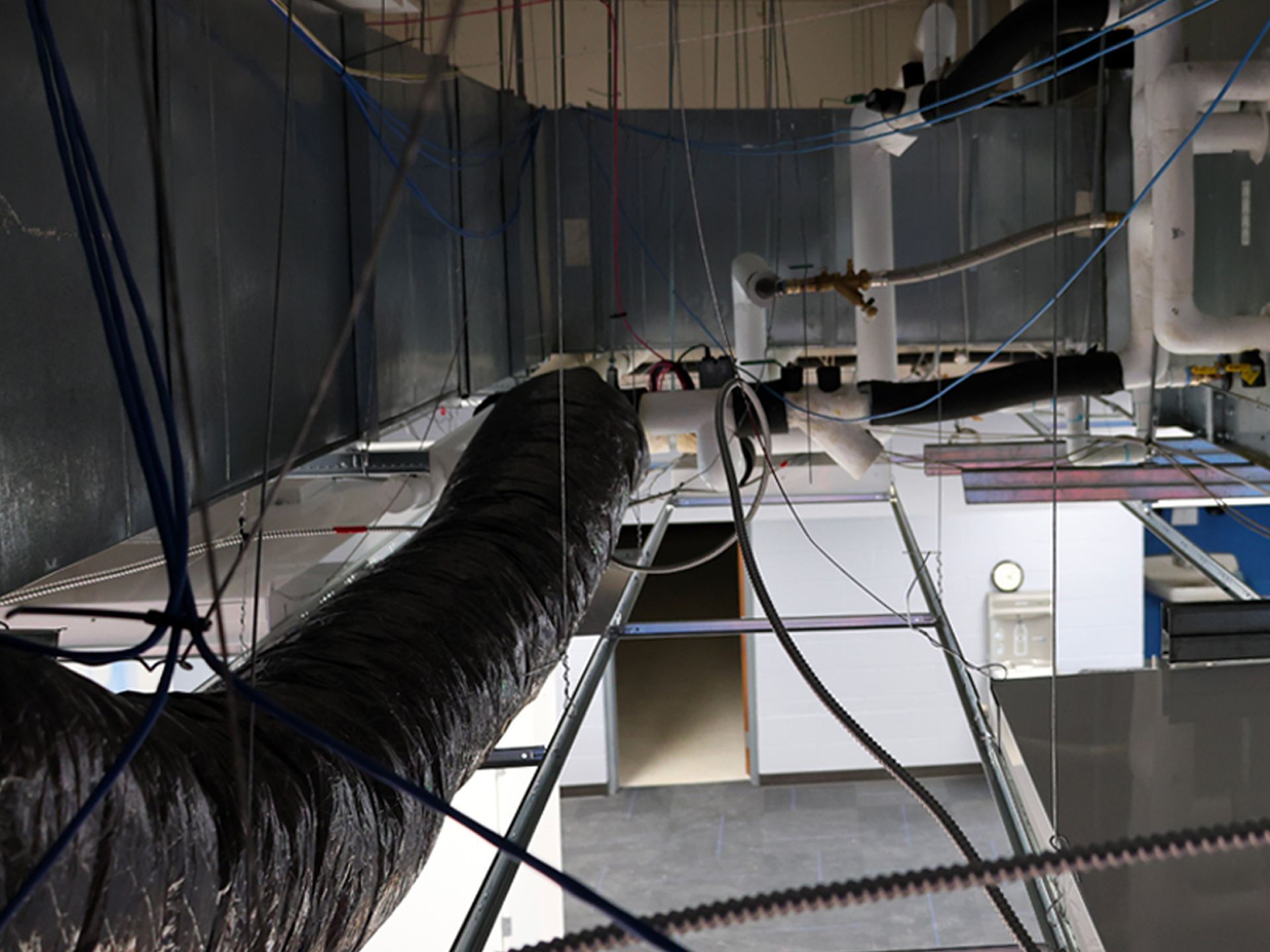 A view of ceiling infrastructure including silver ducting, pipes, wires, and suspension cables in an unfinished room.