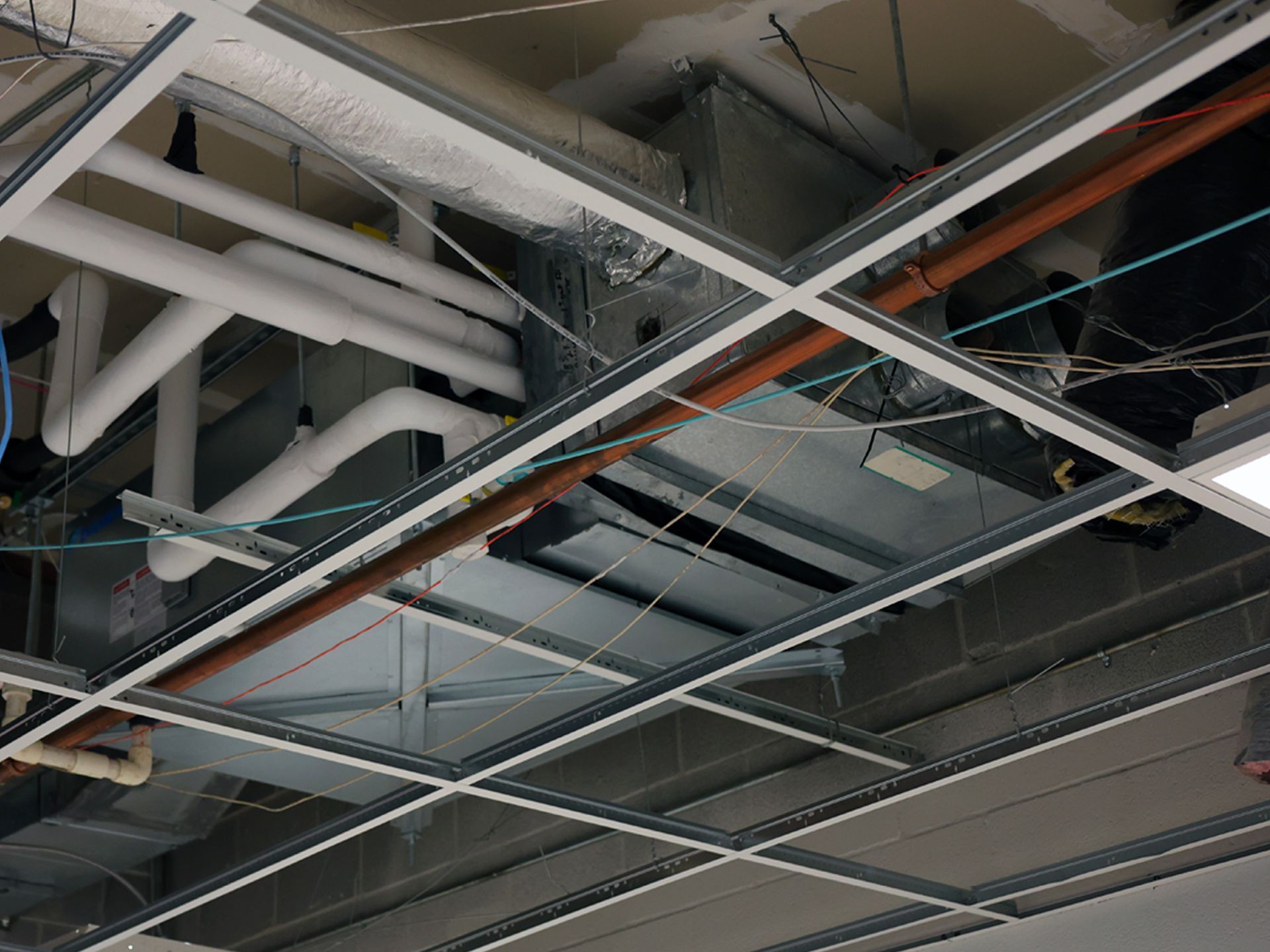 Unfinished drop ceiling showing exposed metal framing, white piping, ductwork, and various wires in a commercial space.