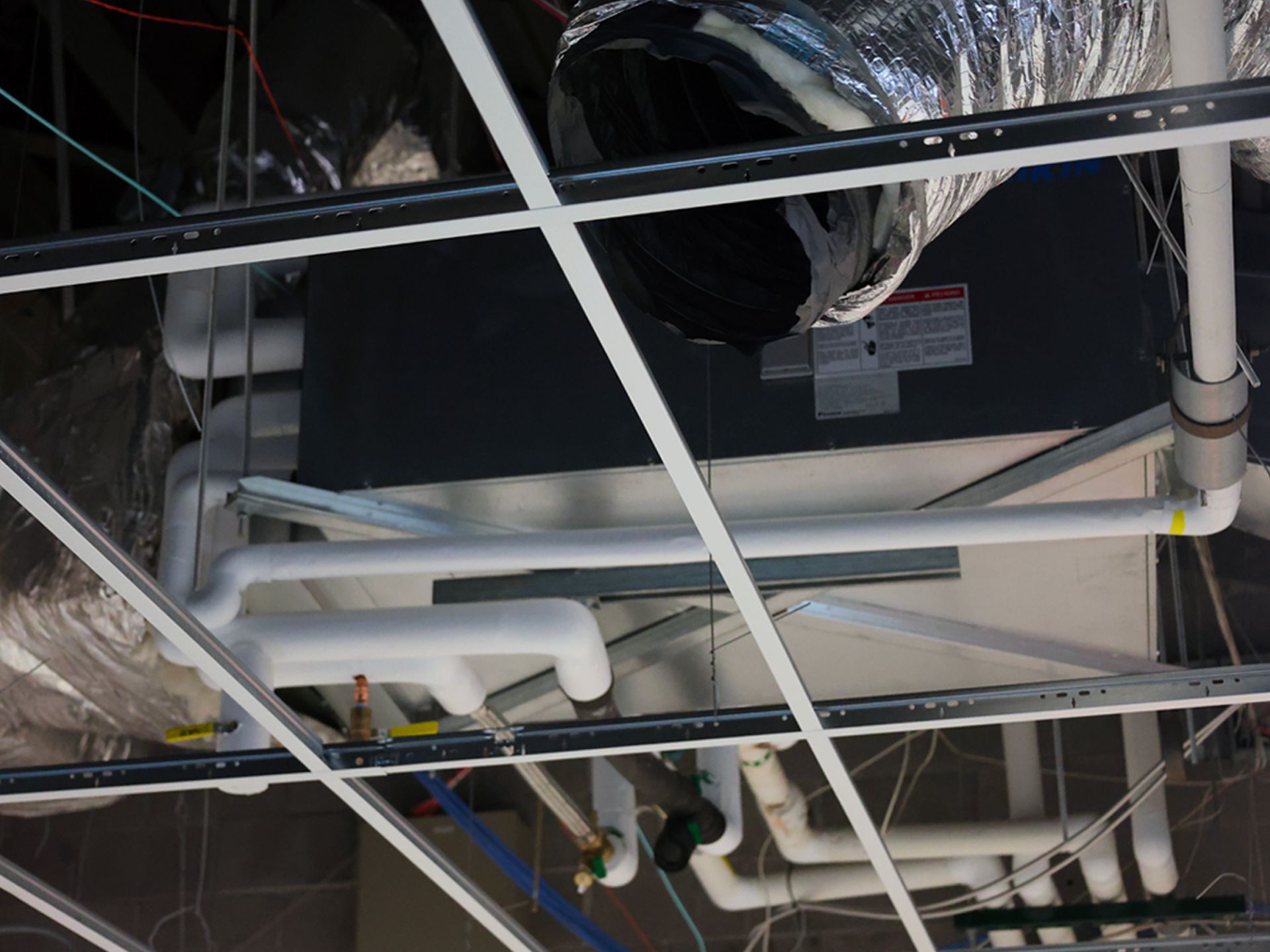 A ceiling-mounted HVAC unit with white insulated pipes and ductwork suspended above a metal grid frame.