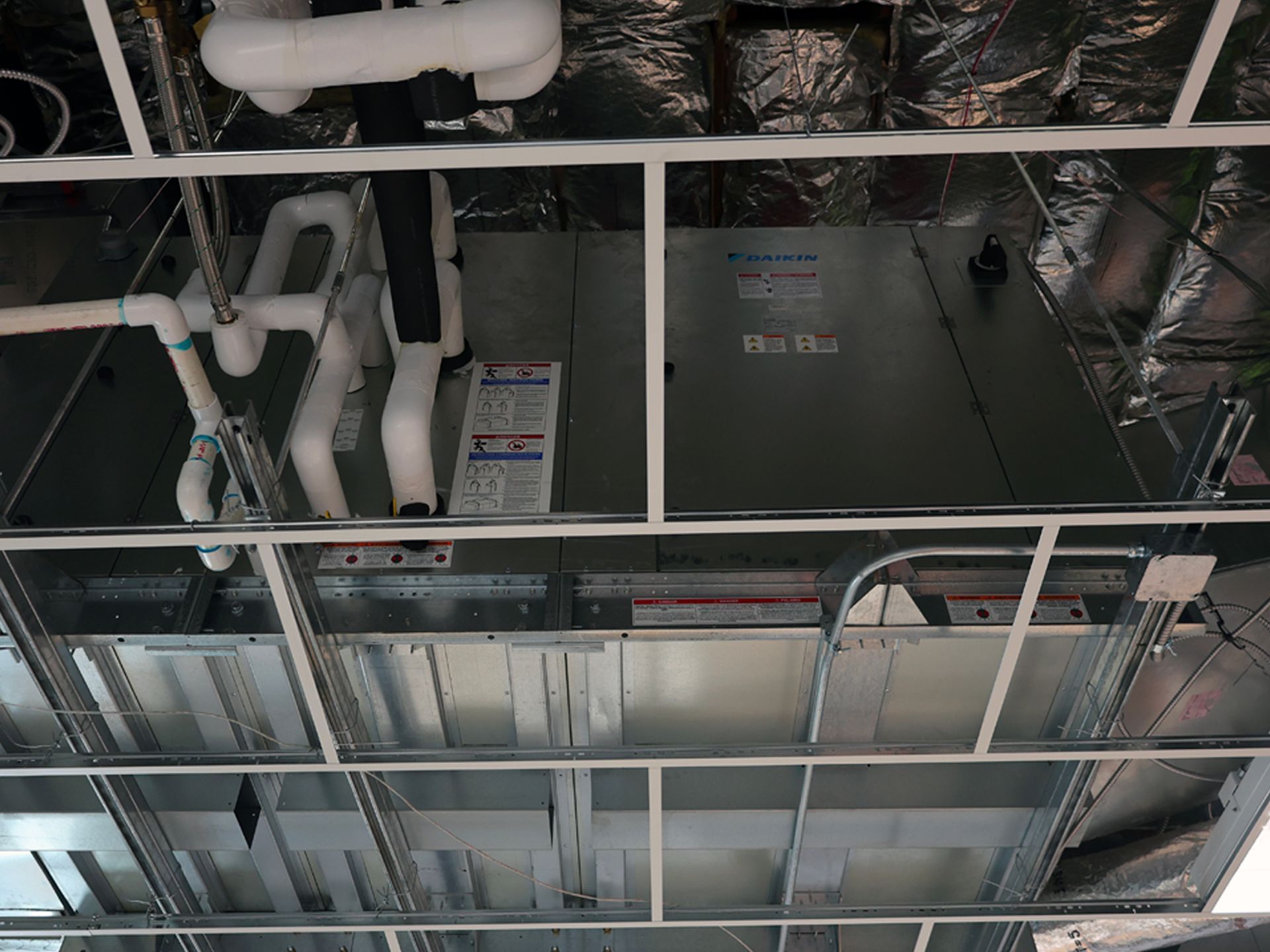 An HVAC air handler unit installed above a suspended ceiling grid with white pipes and silver insulation.