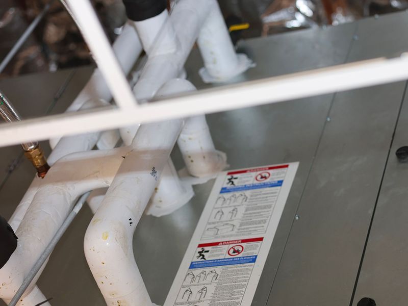 White PVC plumbing pipes attached to a metallic HVAC unit with a caution label.